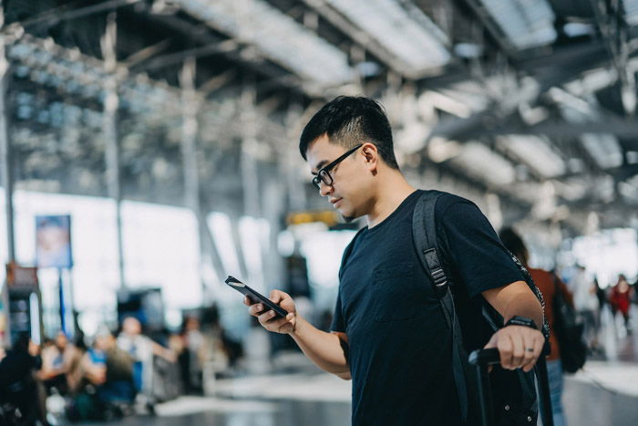Young Asian male backpacker with suitcase checking on smartphone while walking in the international airport hall