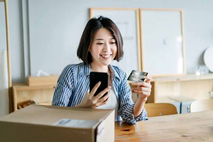 Cheerful young Asian woman shopping online with smartphone and making mobile payment with credit card on hand, receiving a parcel by home delivery service. Technology makes life so much easier