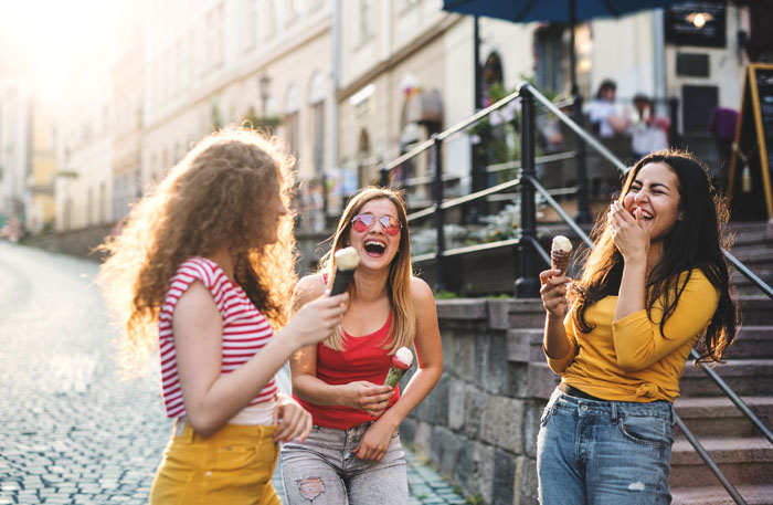 Three cheerful female teenager friends with ice cream standing on the street, laughing.